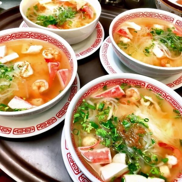 Four bowls of Vietnamese pho with shrimp, crab sticks, and herbs on a tray.