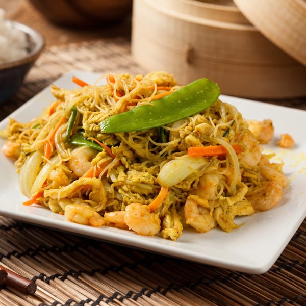 Curry shrimp and chicken Singapore noodles with snow peas, carrots, and onions on a white plate.
