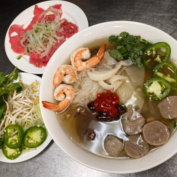 A large bowl of pho with shrimp, meatballs, tripe, and chili sauce, served with sides of raw beef and bean sprouts.